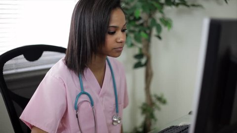 A pretty young African American nurse or nursing student working at a computer answers a phone call. Camera pans and tilts slightly, then tilts down to a the keyboard and back up to the girls face.