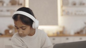Cute teenage girl wearing wireless headphones while sitting at desk and typing on modern laptop. Young female spending time at home for education.