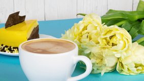 Fresh aromatic espresso coffee with froth. Cup of coffee with a bouquet of yellow tulips and a slice of cake on a light blue background with copy space. Breakfast for a loved one, 4k
