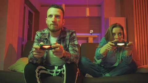 Emotional diverse gamers are holding joysticks and competition in intense video game on gaming console. The woman is winning the game. Cozy room is lit with warm and neon light.