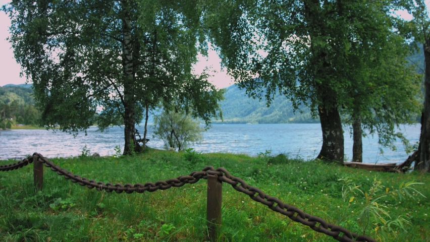 On the shore of the lake there is a massive chain in the form of a fence. It hangs on posts dug into the ground. The shore is covered with grass and trees grow on it.