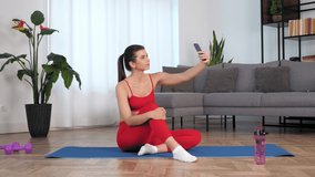 Smiling sportive woman fitness blogger sitting on yoga mat listen tells online video call on selfie camera smartphone. Positive fit sports girl records stories after workout exercise training at home
