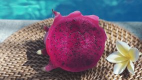 Fresh red dragon fruit cut into two halves with a spoon stuck into the pulp on the edge of bubbling blue swimming pool with white tropical flowers frangipani, moving shade from palm branch in Bali