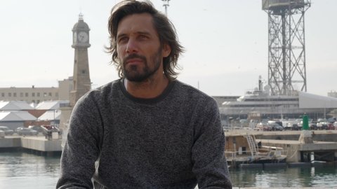 Handsome bearded man sits and sips hot drink with Barcelona Port Vell harbor in background. Spain