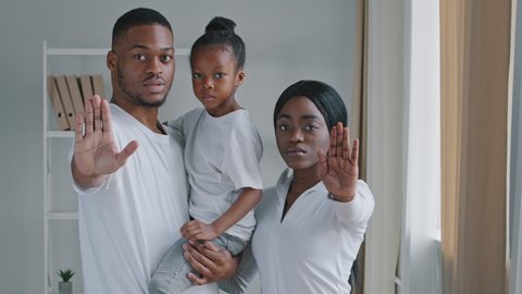Serious afro american family parents with little daughter child kid girl standing looking at camera putting palms in front of them making refusal prohibition disagreement stop gesture, keep distance