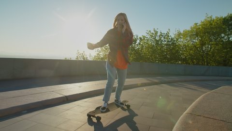 Active cool skillful attractive young woman skateboarder in trendy clothes talking on smartphone, skateboarding on city street in early morning during beautiful sunrise while enjoying leisure outdoors