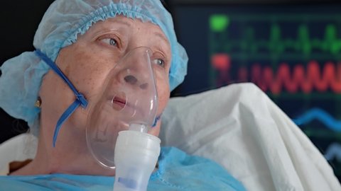 Portrait of old woman in oxygen mask on hospital bed against background of monitor with vital signs