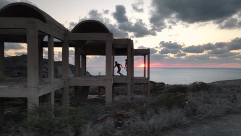 Aerial dolly left shot of two men running and parkouring inside a construction on the coast at sunset