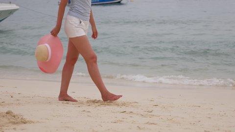 Woman walk along sea beach, dangle hand with pink summer hat, tracking shot. Tourist lady leisurely stroll at shore of sea bay, spend slow vacation days at tropical country