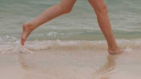 Woman run barefoot at swashing waves on sea beach, slow motion low half shot. Tracking camera show only legs of happy and carefree tourist. Small waves move towards and break, water splash around feet