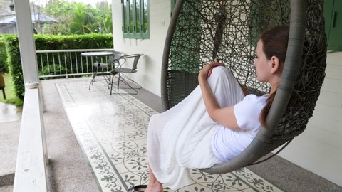 Woman sit down and relax in hanging cocoon chair, look to green garden, spend rain hour at villa porch. Wide angle view from behind, round swinging seat slightly sway