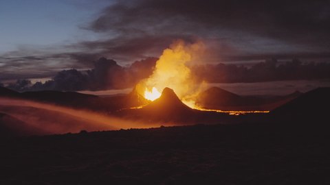 A long-distance shot from the view of a rugged hill of an erupting volcano crater emitting golden-colored lava and cloud-like smoke on the backdrop of a stunning faded purple sky.