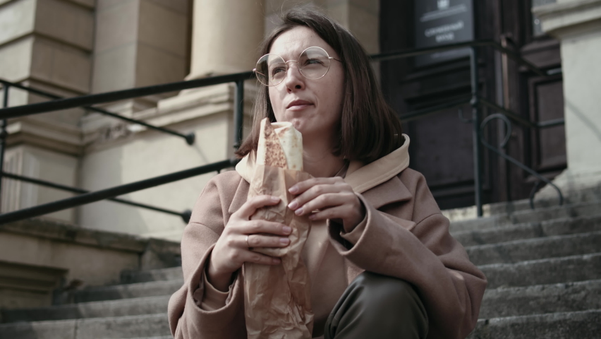Closeup of young female wearing stylish outwear sit outdoors in city streets and eat street food and drinks tea or coffee to go. Woman in beige coat using has a lunch with street food