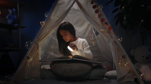 Curious teen with long black hair in makeshift hut looking to book at home in evening. Young girl sitting on floor and using hand flashlight. Concept of leisure and free time