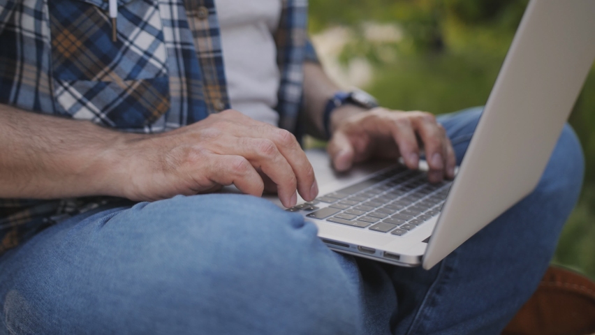 Close-up of the hands of a middle-aged man working on a laptop in an outdoor park.