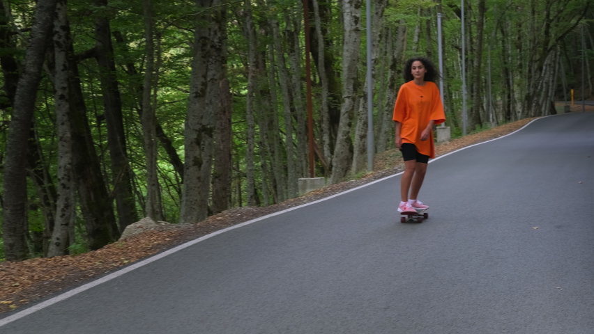 Slow motion shot of a pretty young brunette in orange oversized t-shirt skateboarding down the road running through the scenic wood. Enjoyable active leisure