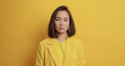 Serious young Asian woman with dark hair looks directly at camera dressed in formal clothes smiles positively isolated over yellow background. Elegant female model prepares for business meeting