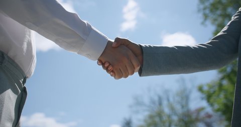 International partnership. Close up outdoor shot of caucasian businessman and african american business lady shaking hands, confirming cooperation and agreement over blue sky background, slow motion