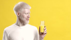 Happy Albino Guy Holding Glass Of Orange Juice Posing Smiling To Camera Standing Over Yellow Studio Background. Healthy Drink, Vitamins And Hydration Concept. Copy Space