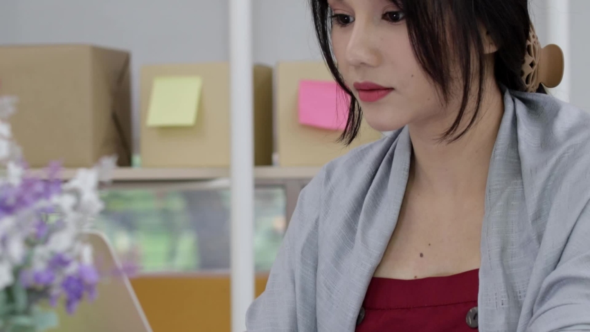 Busy asian woman red top hair tied typing address quickly on laptop sending package for ecommerce while looking at notebook and writing with pencil. Panning from face to object.