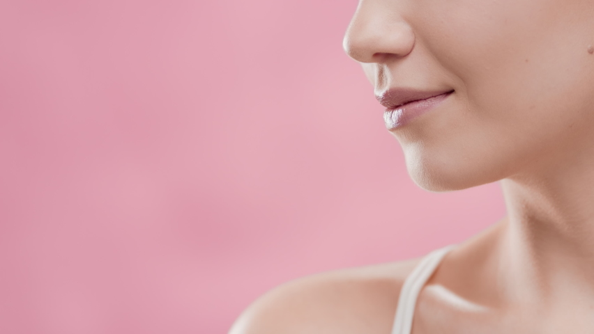 Close-up beauty portrait of young fit cute blonde woman in white bikini turns head to the camera touching her jawline and smiling wide against pink ripple background | Face care cosmetics commercial