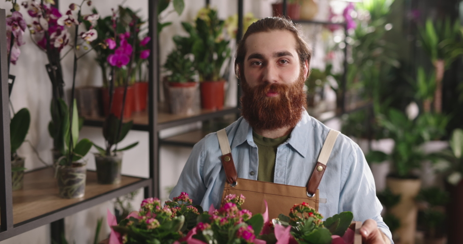 Hipster millennial entrepreneur working in flower shop, taking care of plants. Male florist looking at camera - small business people 4k footage portrait