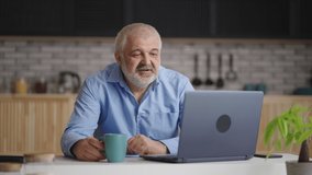 middle-aged man is chatting by video call by laptop, sitting alone at home kitchen, drinking tea