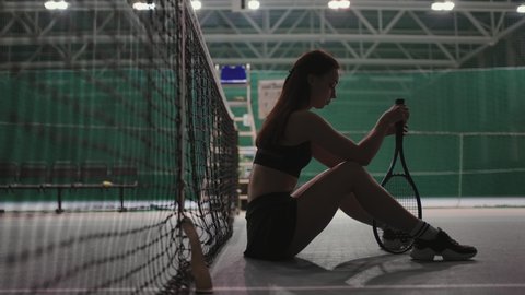 pensive female tennis player is sitting on floor on tennis court after game or training, twisting racquet