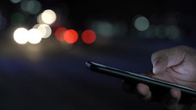 Close-up of a Hand with a Smartphone on the Background of Night City Lights and Headlights Passing by Cars. Nightlife Search Concept. Blurred Video. Film Grain
