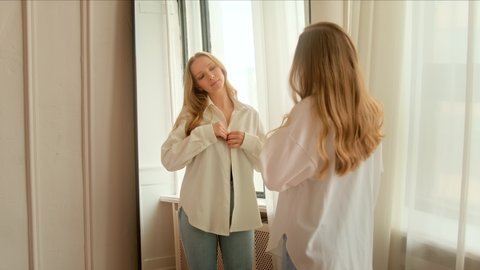 Young woman getting dressed looking in mirror. Trying on clothes enjoying preens herself, admires and straightening her clothes and getting ready for date. 