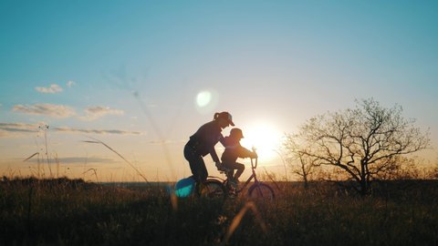 Silhouette of mother teaching little son to ride a bike at meadow during sunset. The kid turns the bike close-up. Caring young mother teaches her son to ride bicycle. Concept of friendly family.