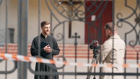 a young priest gives an interview on camera to the churchyard