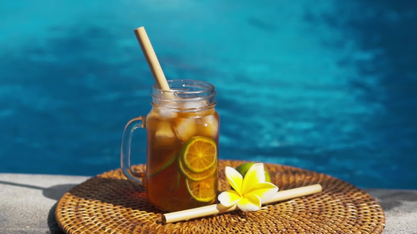 Video footage of glass mason jar with iced tea, cold with ice cubes and slices of lime, bamboo straw, whole lime, frangipani flower and bubbling blue swimming pool on background.
 | Shutterstock HD Video #1073204216