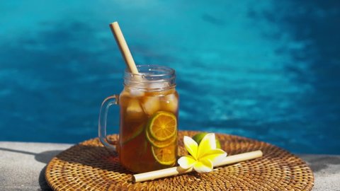 Video footage of glass mason jar with iced tea, cold with ice cubes and slices of lime, bamboo straw, whole lime, frangipani flower and bubbling blue swimming pool on background.
