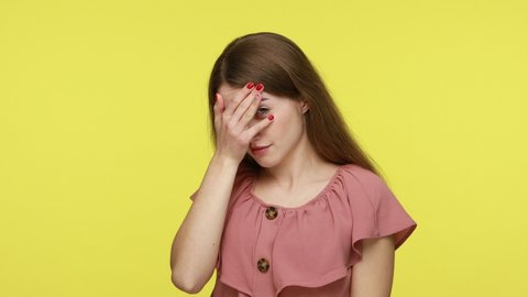Curious brown haired woman in dress covering eyes with palm of hand, peeking through fingers, looking with suspicion, shy to watch forbidden event, spying. Indoor shot isolated on yellow background.