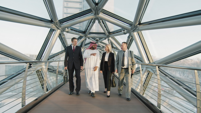 Front-view full-shot slowmo of team of Asian, Arab and Caucasian male and female business partners talking while walking along indoor glass bridge of modern high-class office building Royalty-Free Stock Footage #1073204405