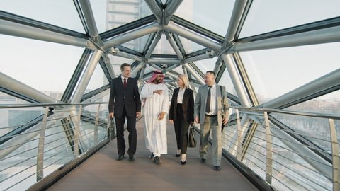 Front-view full-shot slowmo of team of Asian, Arab and Caucasian male and female business partners talking while walking along indoor glass bridge of modern high-class office building