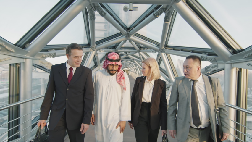 Dolly-out front-view medium slowmo of successful multi-ethnic team of male and female middle-aged business partners talking while walking along indoor glass bridge of modern high-class office building