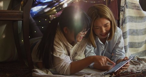 Asian mother and daughter smiling while using digital tablet under blanket fort at home. motherhood, love and togetherness concept