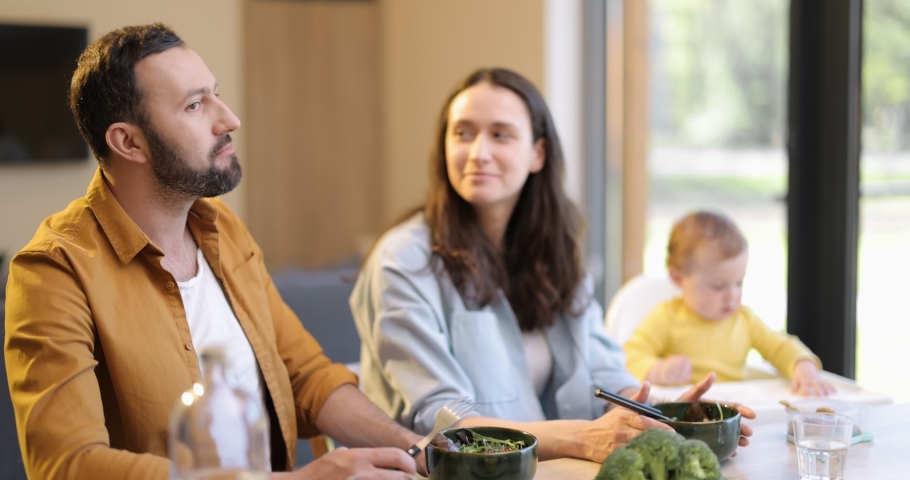 Young happy family with a one year baby boy during a lunch time at home. Concept of healthy vegan eating and happy parenting. Man kiss a wife