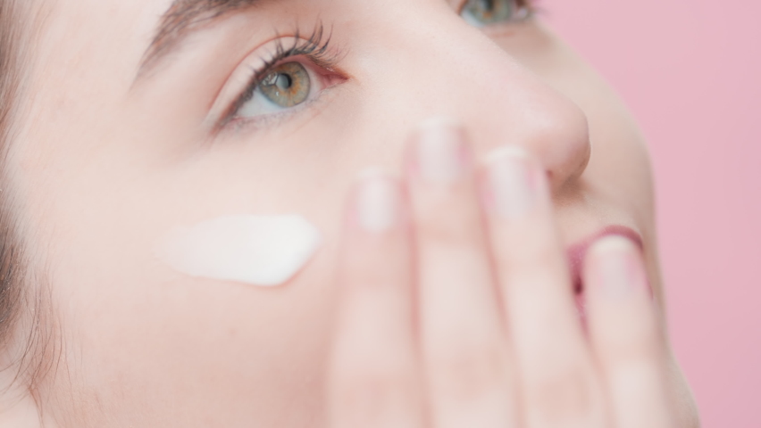 Extreme close-up of young good-looking slim European skincare woman applies moisturizer under her eye smiling wide against pink ripple background | Moisturizer applying on shot for skincare commercial | Shutterstock HD Video #1073216837