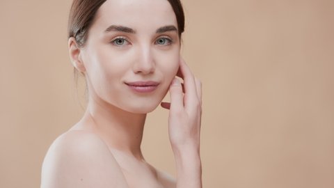 Gorgeous fit Caucasian woman with dark hair and naked shoulders touches her jawline with hand turning body, looking at the camera and smiling wide against beige ripple background | Skin care concept