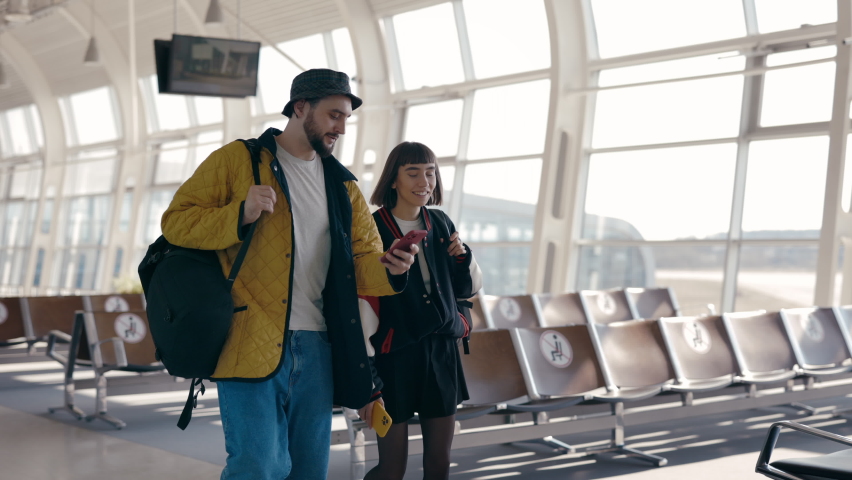 Happy couple of travellers with backpack walking together at airport lounge, using smartphones and talking. Young man and woman in stylish outfits travelling abroad. Royalty-Free Stock Footage #1073223524
