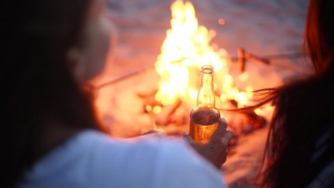 Young woman drinking beer of glass bottle sitting around bonfire with friendsand fryin sausages on sandy beach. Girl with beverage relaxing at campfire in the dusk. Female chilling with lemonade.