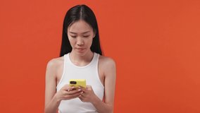 Young asian woman 20s years old wears white tank top shirt get video call using mobile cell phone doing selfie talk conducting pleasant conversation greet with hand on orange color background studio