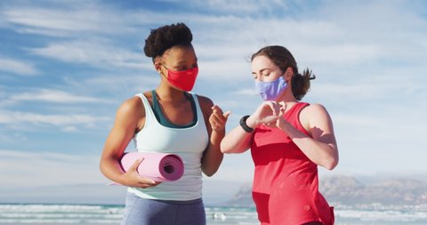 Two diverse female friends wearing face masks holding yoga mats at the beach. healthy active lifestyle, outdoor fitness and wellbeing.