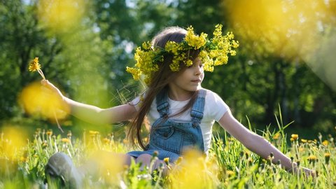 Little cute girl in a flower wreath on her head collects dandelions while sitting on a sunny meadow
