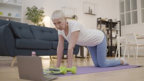 Mature lady learning to do yoga pose at home, watching tutorial video on laptop