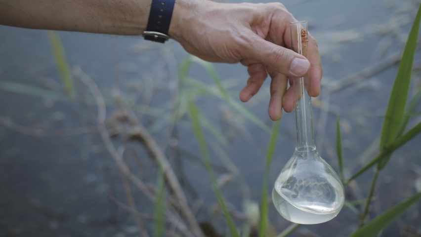 Men's Hand draws water from a polluted reservoir and holding test tube for analyzes with water on the background of the reservoir, the concept of water purity, | Shutterstock HD Video #1073233760