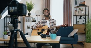 Blogging concept where attractive emotional happy smiling young african american man holding online performnace for internet audience while playing on the guitar at home